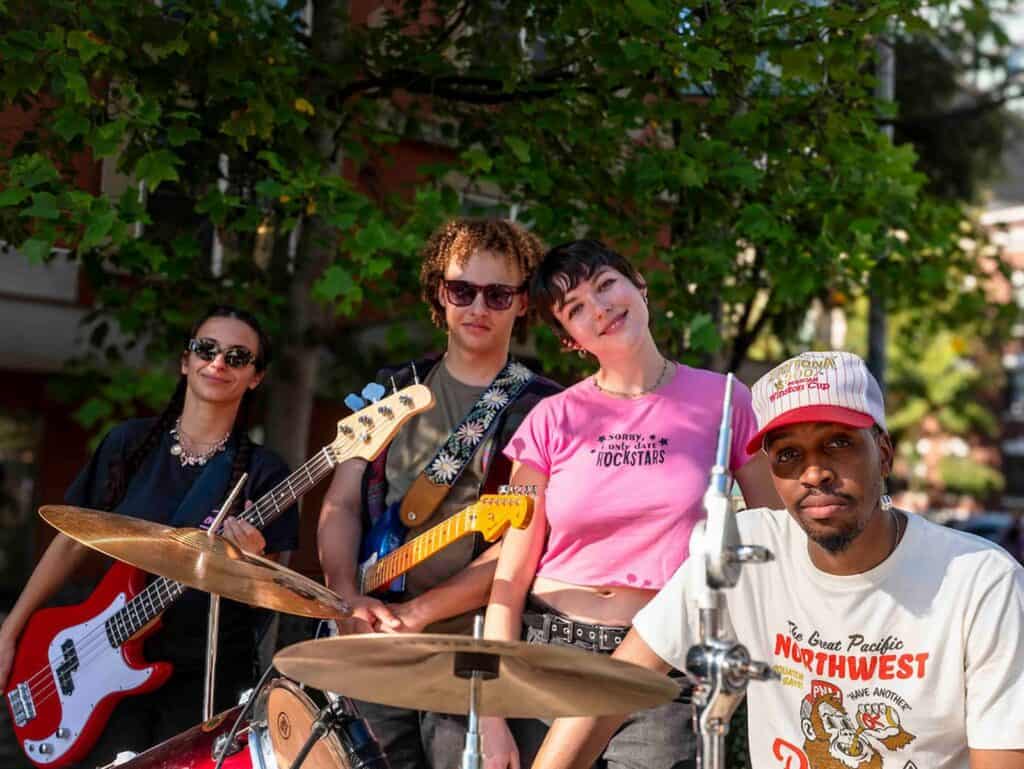Four musicians with instruments stand outdoors in front of trees: one with a bass guitar, one with an electric guitar, one at the drums, and one standing in front.
