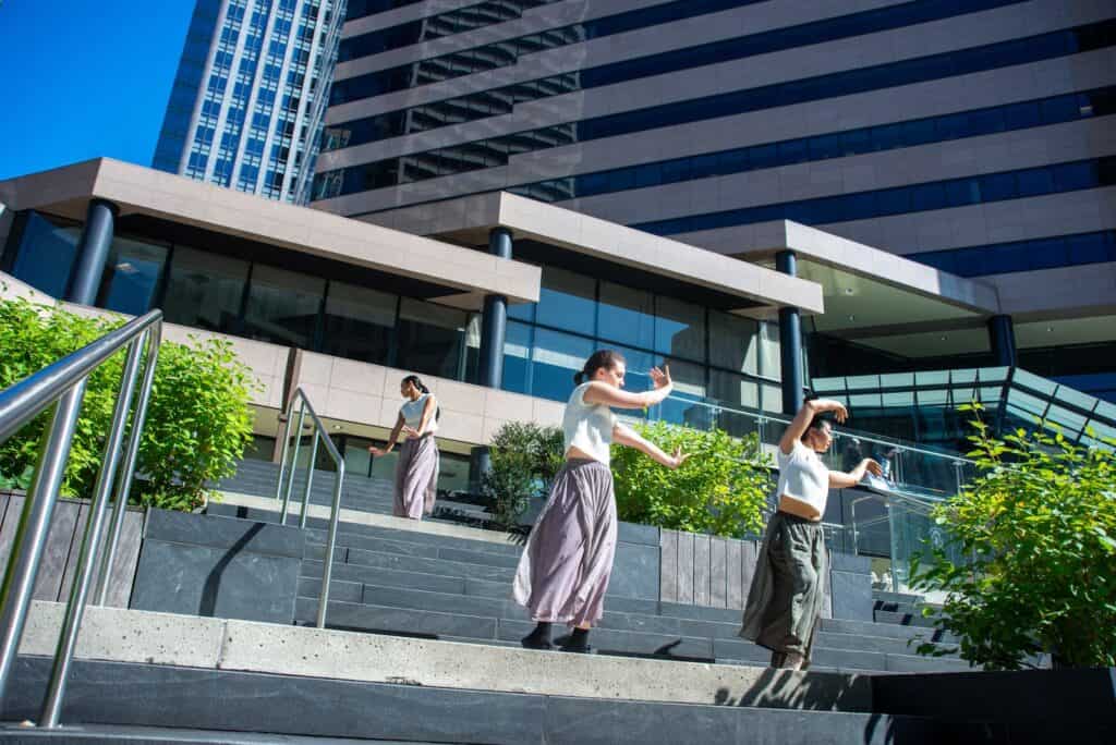 Three women stand and gesture on outdoor steps in front of a modern office building in Downtown Seattle, with greenery and bright sunlight—a lively scene that could be part of an art show or the energetic WALKDONTRUN event.