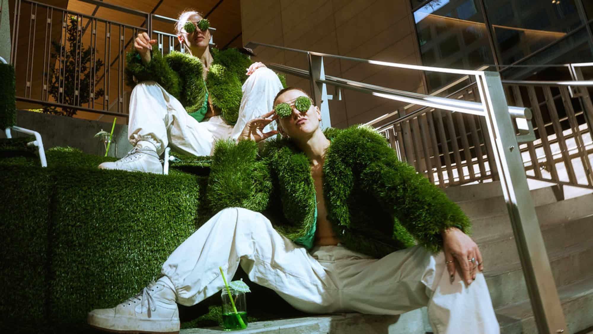 Two people wearing green fuzzy jackets, white pants, and green sunglasses sit on grass-covered stairs, holding green drinks, in a modern indoor setting—an eye-catching scene that could easily be found at an Art Show in downtown Seattle.
