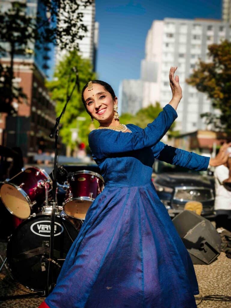 A woman in a blue dress performs a traditional dance outdoors, smiling, with drums and city buildings in the background.