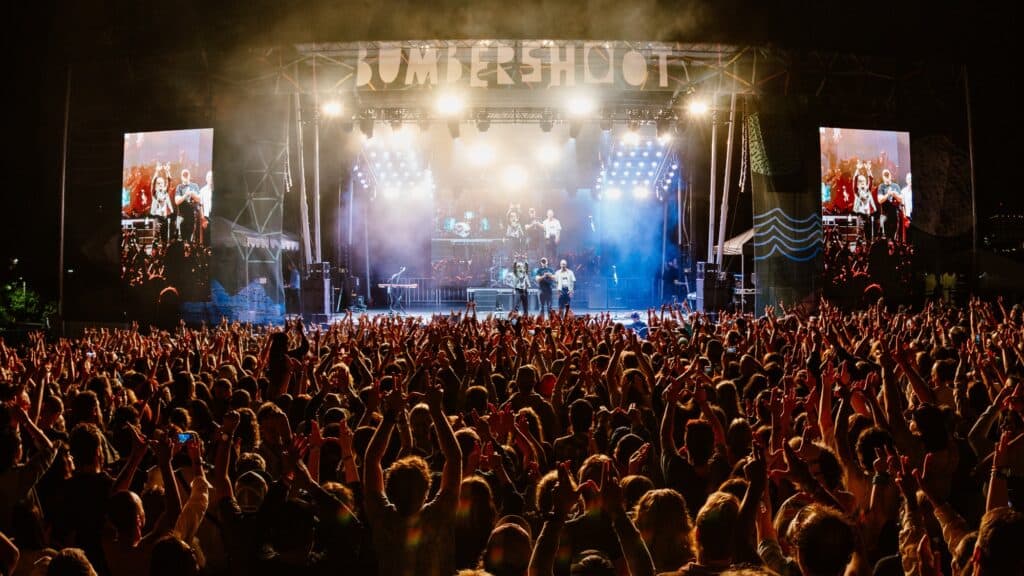 Large crowd watching a band perform on a brightly lit outdoor stage at a music festival, with festival name “Bumbershoot” visible above the stage.