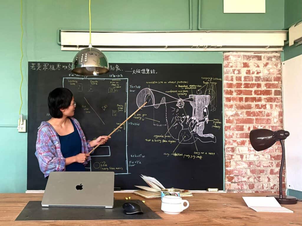 A woman stands by a chalkboard covered in diagrams and text, pointing with a stick. A laptop, papers, and a cup are on the desk in front of her.