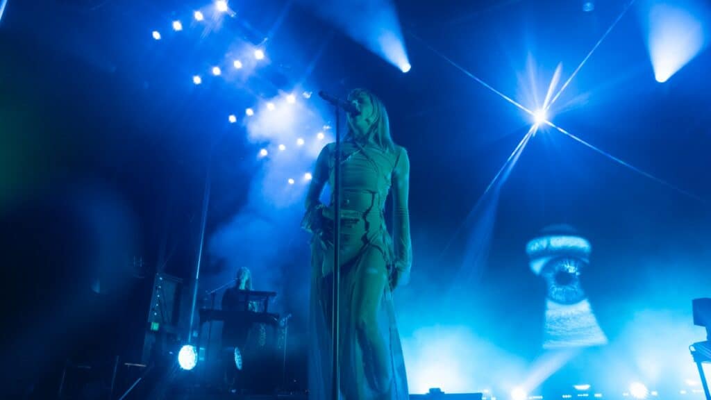 A singer stands at a microphone on a blue-lit stage with dramatic lighting and a keyboard player in the background.