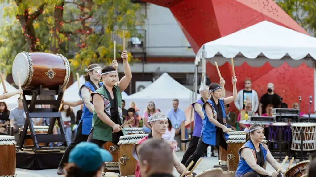 A group of people perform a taiko drumming routine outdoors, wearing colorful vests and headbands, with a large drum and tents in the background.