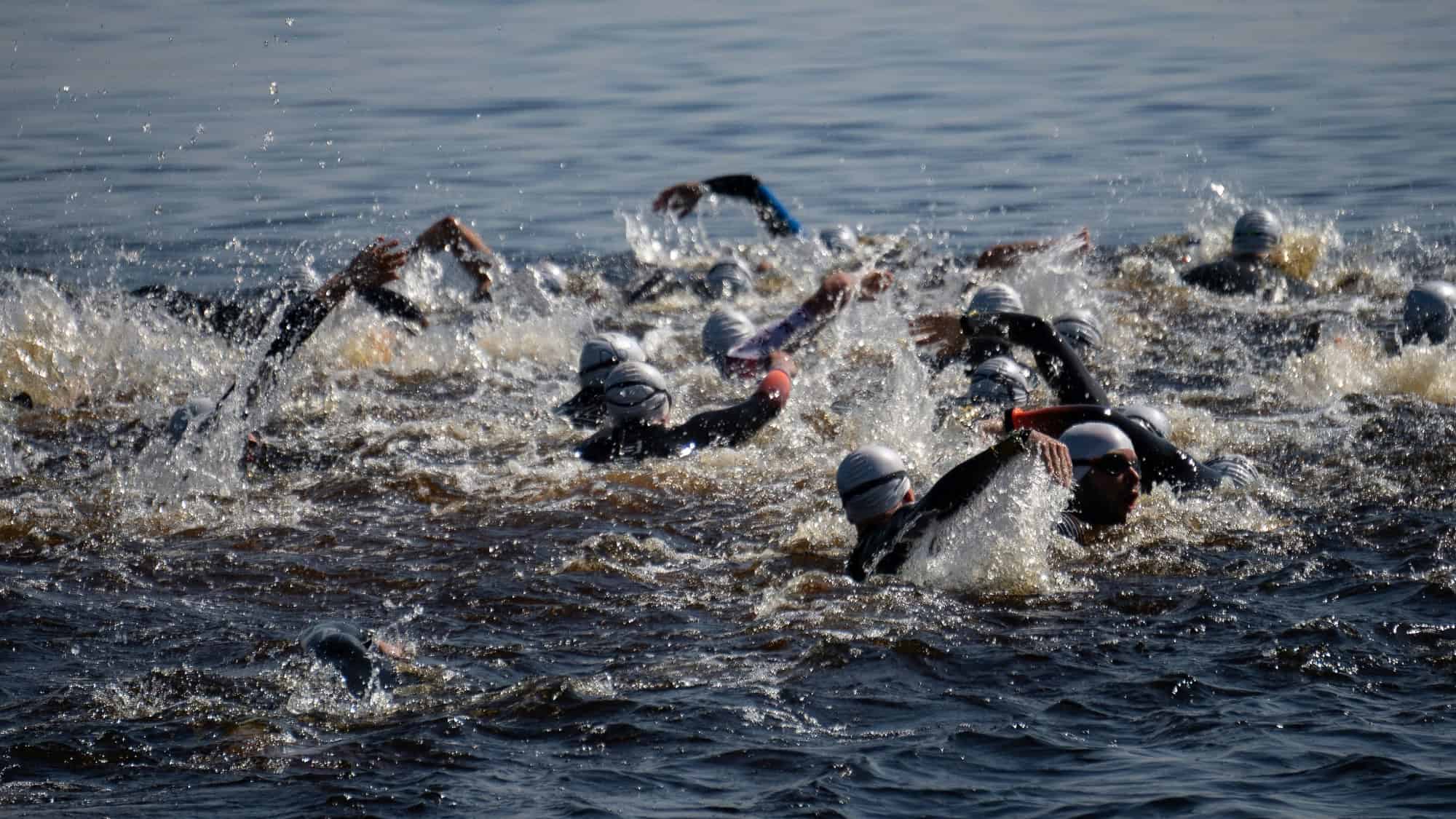 A group of swimmers wearing wetsuits and swim caps compete in open water, creating splashes as they move forward.