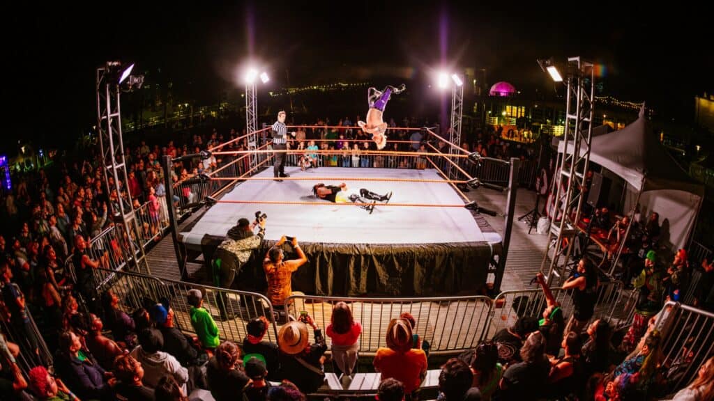 A wrestler performs a backflip from the ropes in a brightly lit outdoor ring at night during the Bumbershoot festival, while a referee and audience watch in amazement.