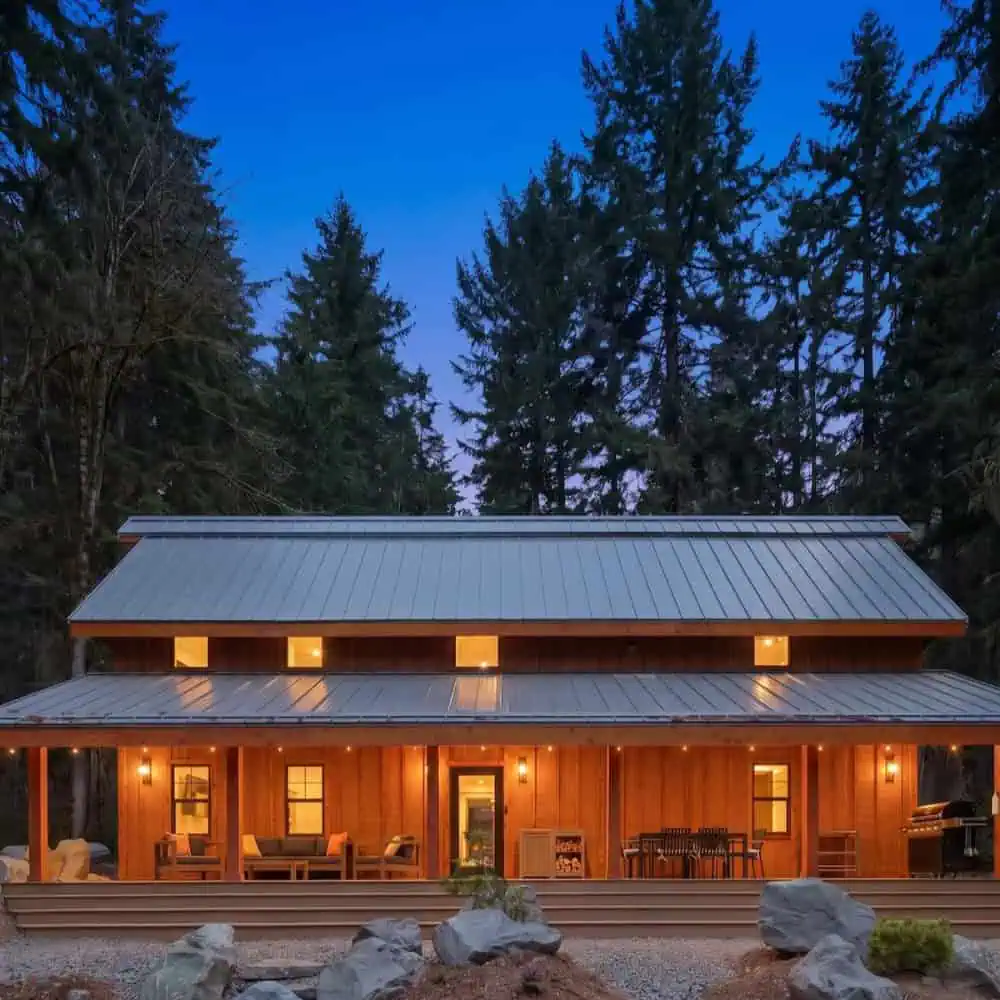 Wooden cabin with a metal roof, warm lights inside, and a covered front porch, surrounded by tall pine trees at dusk.