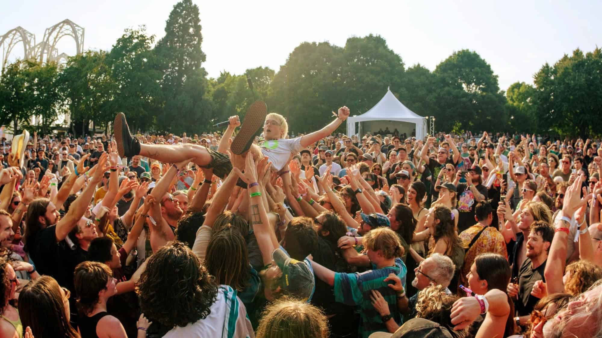A person is crowd surfing above a large group of people at an outdoor music festival with trees and a white tent in the background.