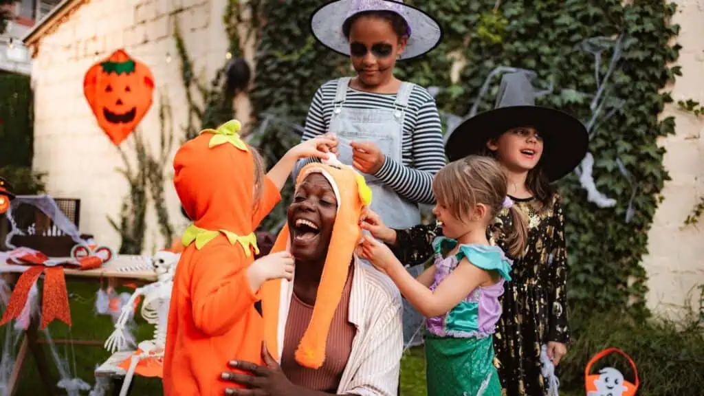 Four children in Halloween costumes from the annual Halloween Costume Hunt help an adult put on a hat outdoors, with festive decorations and greenery in the background.