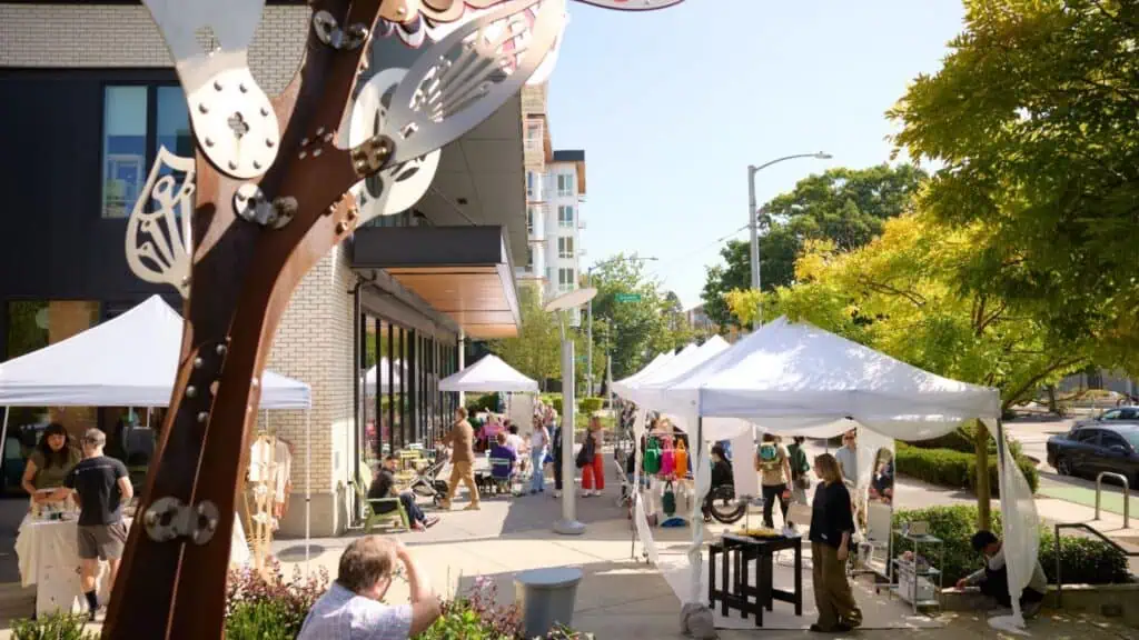 Outdoor art fair with people browsing vendor booths under white tents on a sunny day, with trees and modern buildings in the background.
