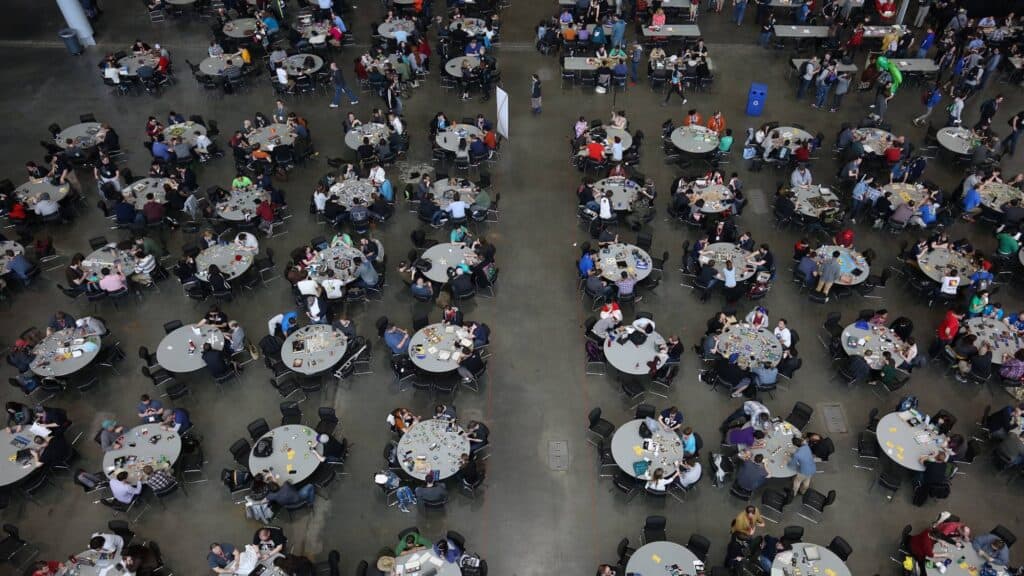 A large crowd of people sit at round tables in a spacious hall at PAX, engaging in conversation and activities while enjoying the magic of shared experiences.