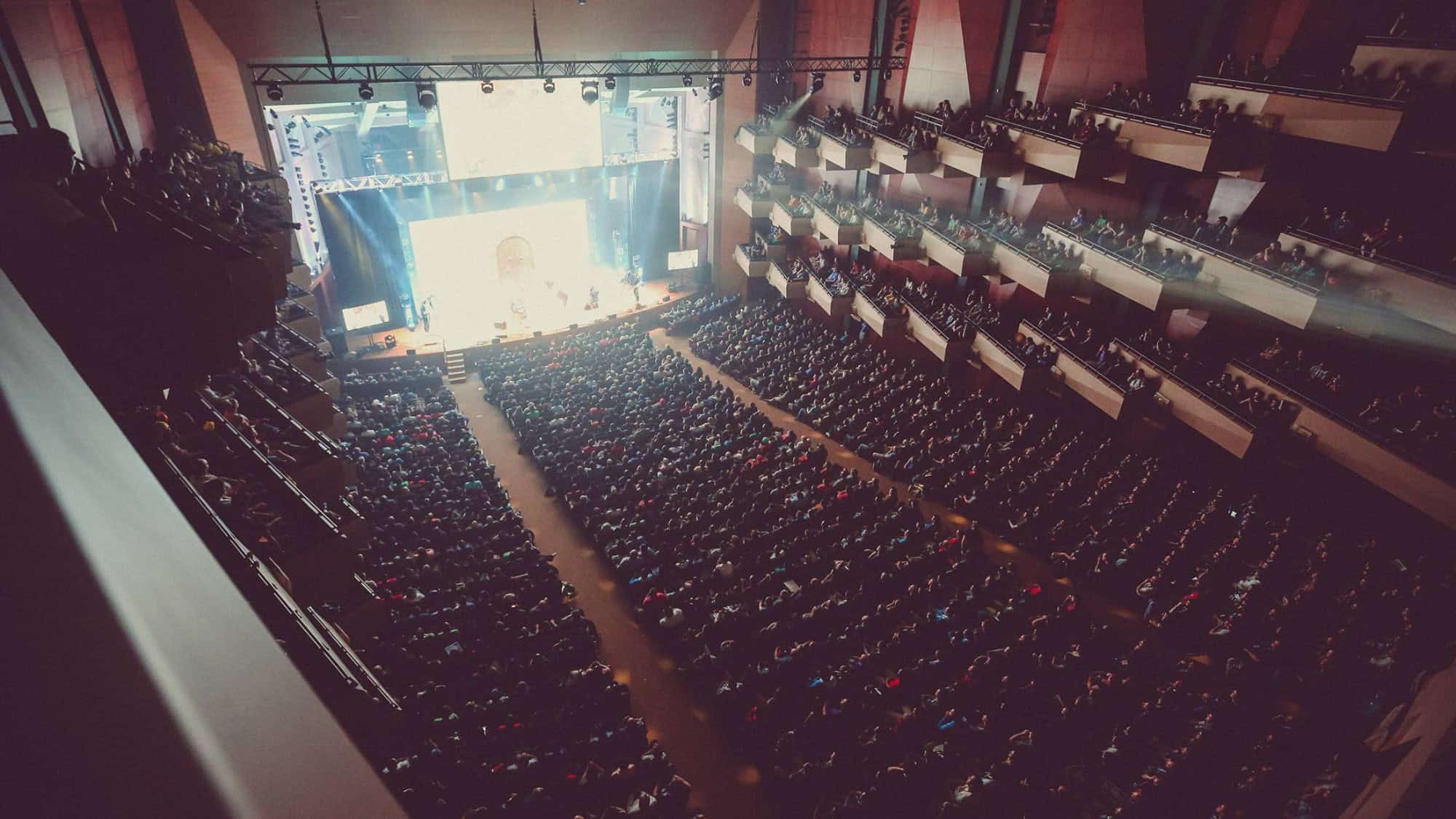 A large audience seated in a modern theater auditorium faces a brightly lit stage during a live performance or event.