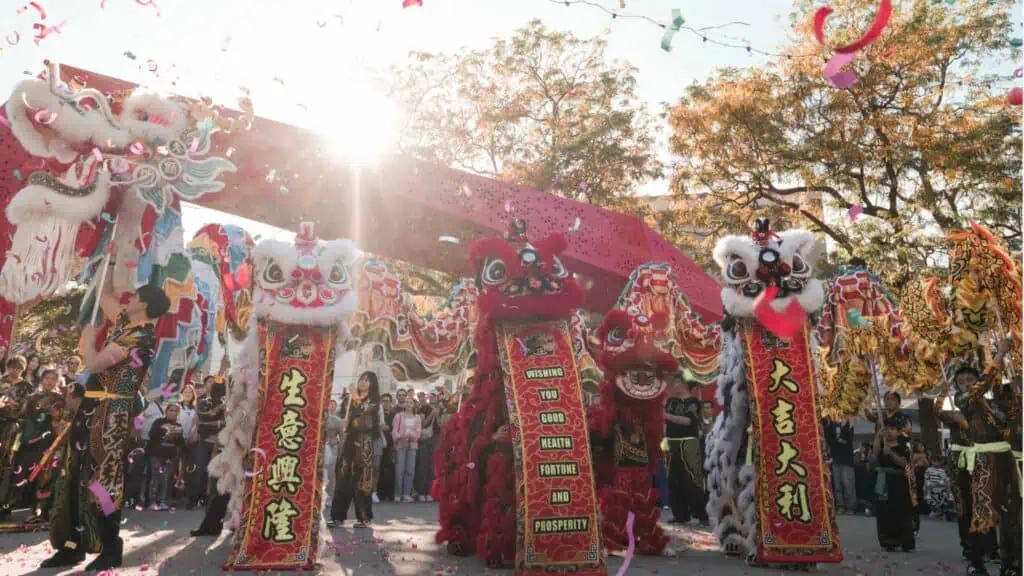 Lion dancers perform outdoors during a festival, surrounded by confetti, banners, and onlookers, with sunlight filtering through trees in the background.