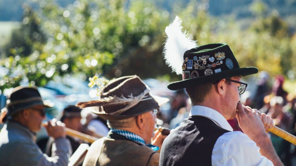 Three people in traditional attire play wooden wind instruments outdoors; one wears a hat decorated with pins and a white feather.