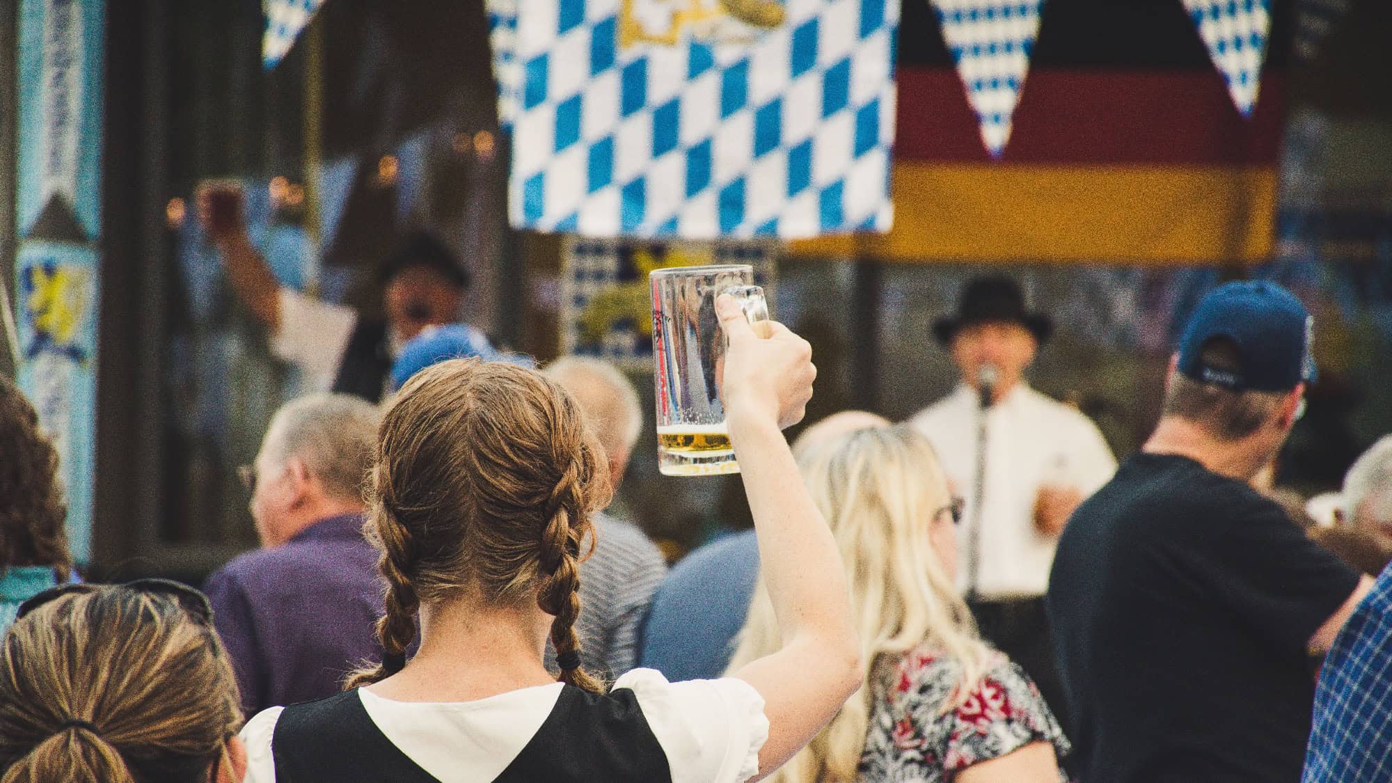 A woman with braided hair raises a beer mug at an outdoor festival, surrounded by a crowd. Blue and white bunting is visible above, and a man speaks into a microphone in the background.