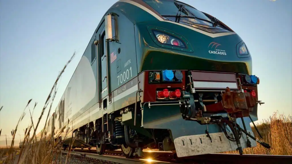 Low-angle view of an Amtrak Cascades passenger train locomotive on railroad tracks, surrounded by dry grass, under a clear sky at sunset.
