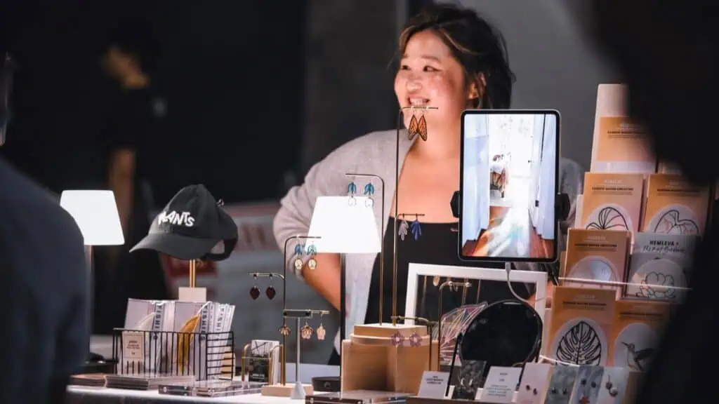 A woman stands behind a table displaying earrings, cards, a hat, and a mirror at an indoor market or craft fair.