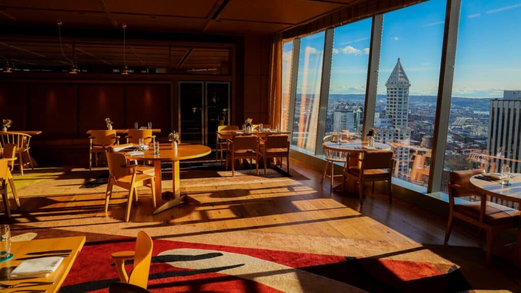 Interior of a luxury lifestyle restaurant with wooden tables and chairs, featuring floor-to-ceiling windows overlooking the Seattle skyline and Smith Tower.