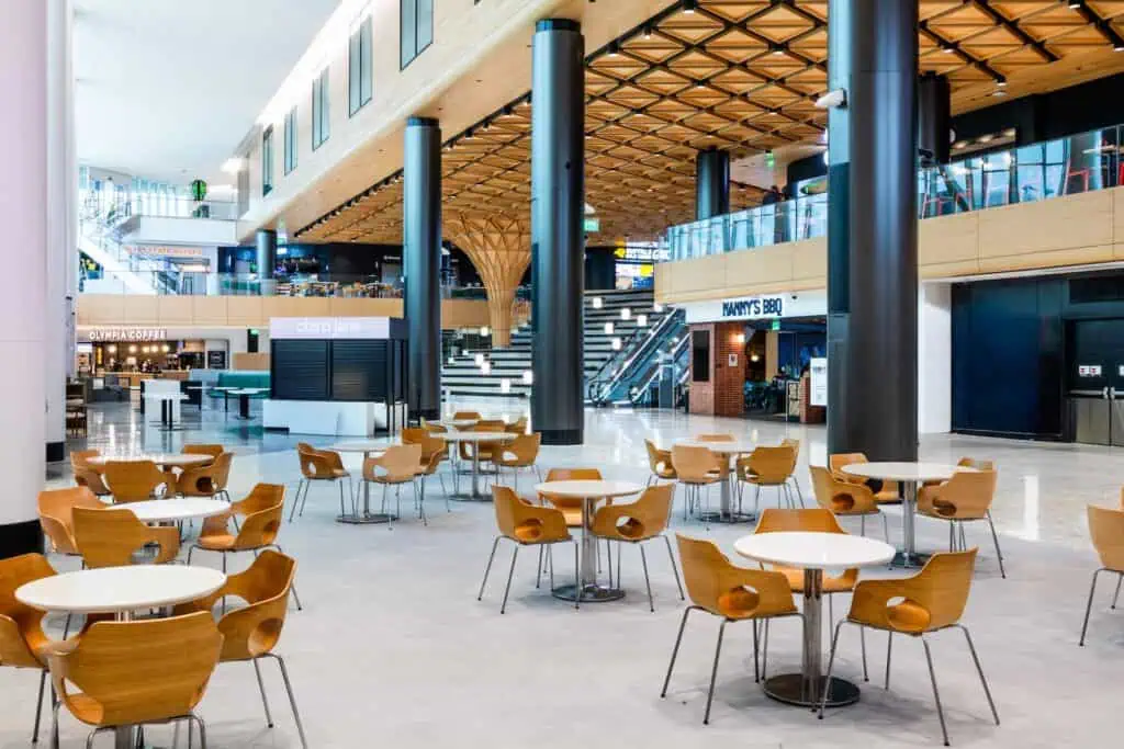 An indoor food court with multiple restaurant storefronts, featuring numerous circular tables surrounded by wooden chairs.