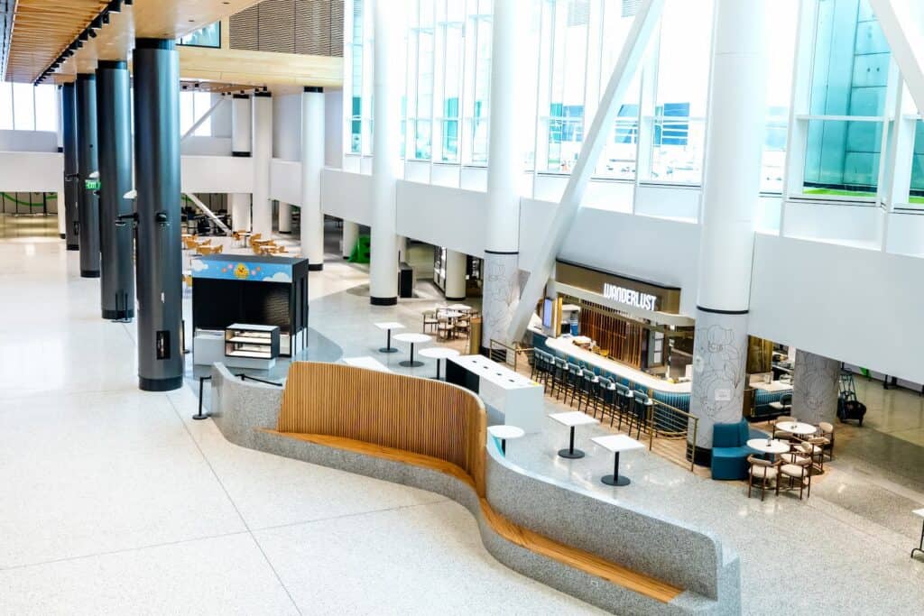 A bright, open airport terminal featuring a lounge area with curved seating and a cafe titled “Wanderlust” with counter stools.