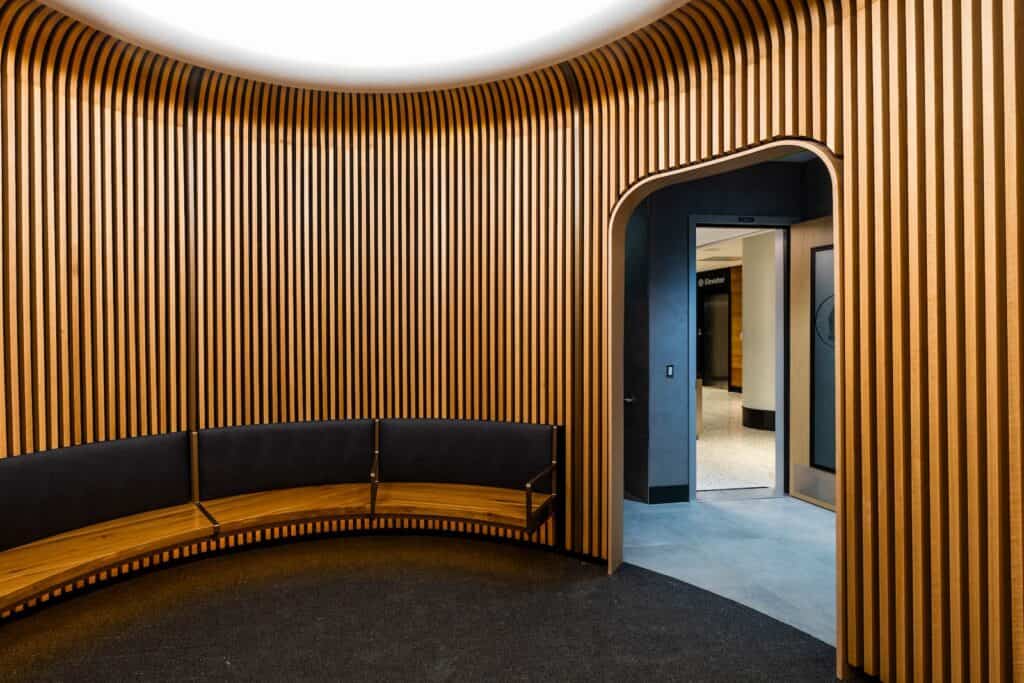 A curved waiting area featuring wood-slatted walls and a dark curved bench, with an arched doorway leading to an elevator lobby.