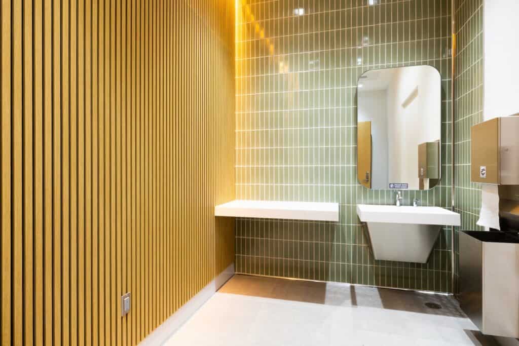 A restroom with a light wood-slatted wall, sage green tile, a floating sink, a mirror, and a wall-mounted paper towel dispenser.