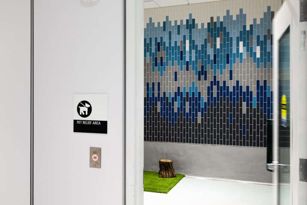 A doorway with a Pet Relief Area sign leads into a room with a blue-and-gray mosaic wall, a patch of artificial grass, and a tree stump.