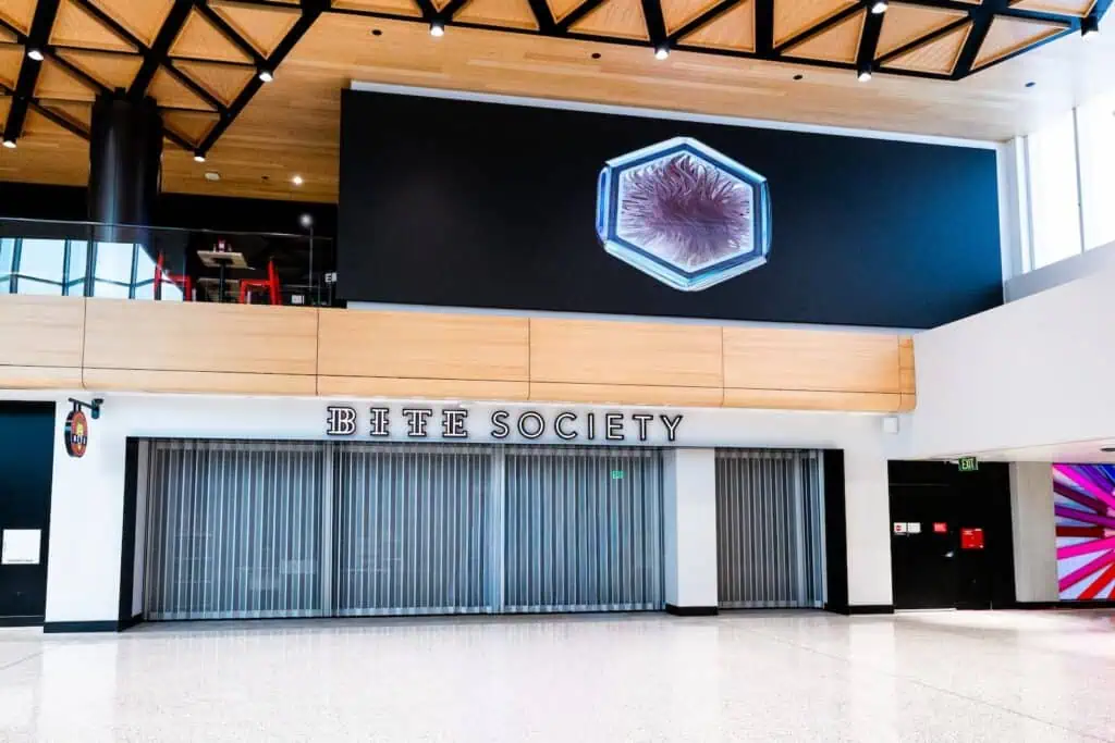 The storefront of Bite Society, featuring closed metal security shutters beneath a wooden header and a large hexagonal wall graphic.
