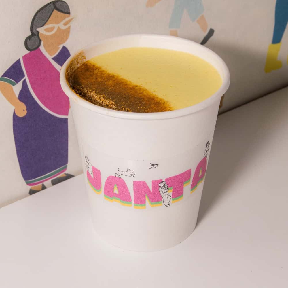 A white paper cup of yellow turmeric latte with a dusting of spice on top, featuring the word “JANTA” in pink letters on a sticker.