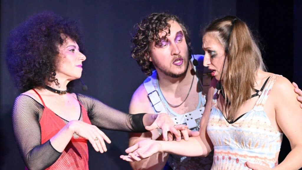 Three performers in theatrical costumes and makeup interacting on stage.