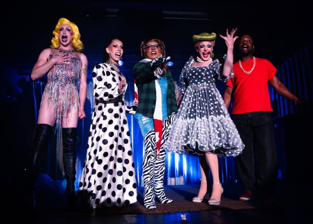 Five performers on stage, four in drag with elaborate makeup and outfits, and one man in a red t-shirt.
