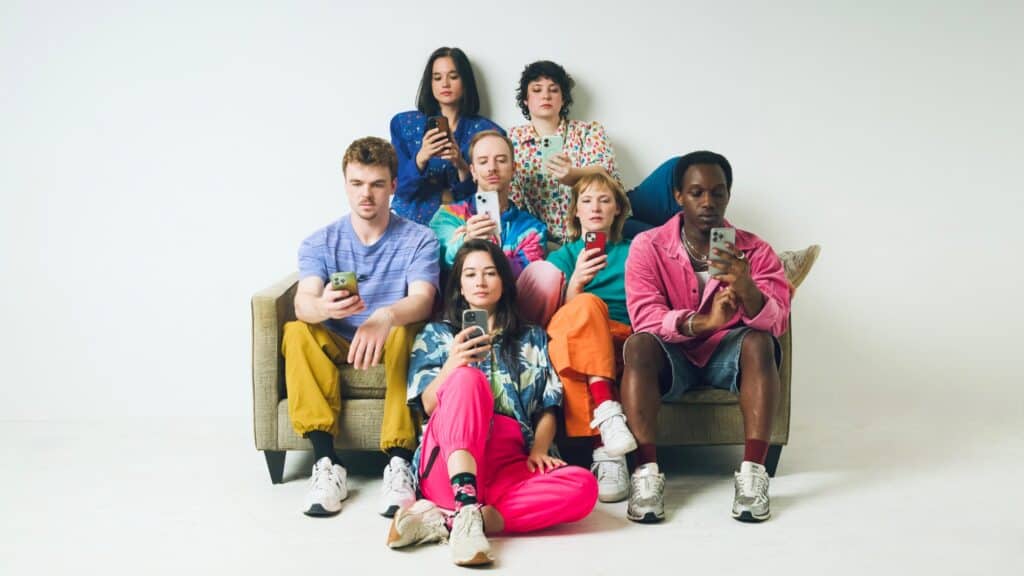 Seven young adults sit on and around a couch, each looking down at their own smartphone in a display of screen addiction.