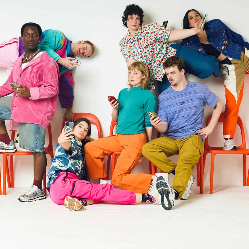 A group of six young adults posing with smartphones against a white wall, some sitting on orange chairs and others leaning or crouching.