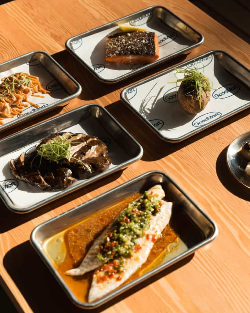 Several small metal trays on a wooden table, each holding a different prepared dish, including salmon, sliced steak, a baked potato, and white fish in sauce.