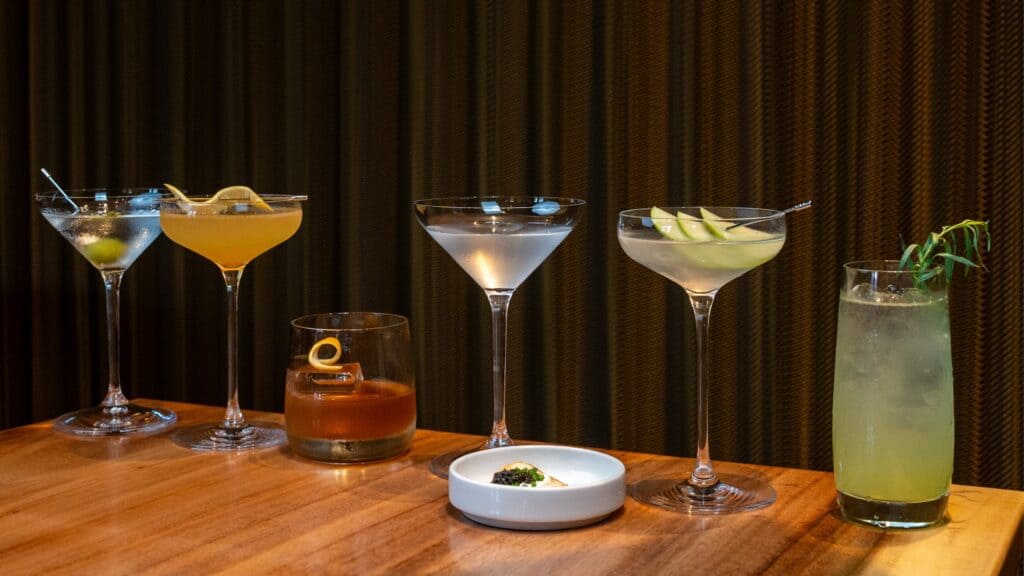 Five cocktails, including a caviar martini, presented in various glassware with a small dish of caviar on a wooden table.