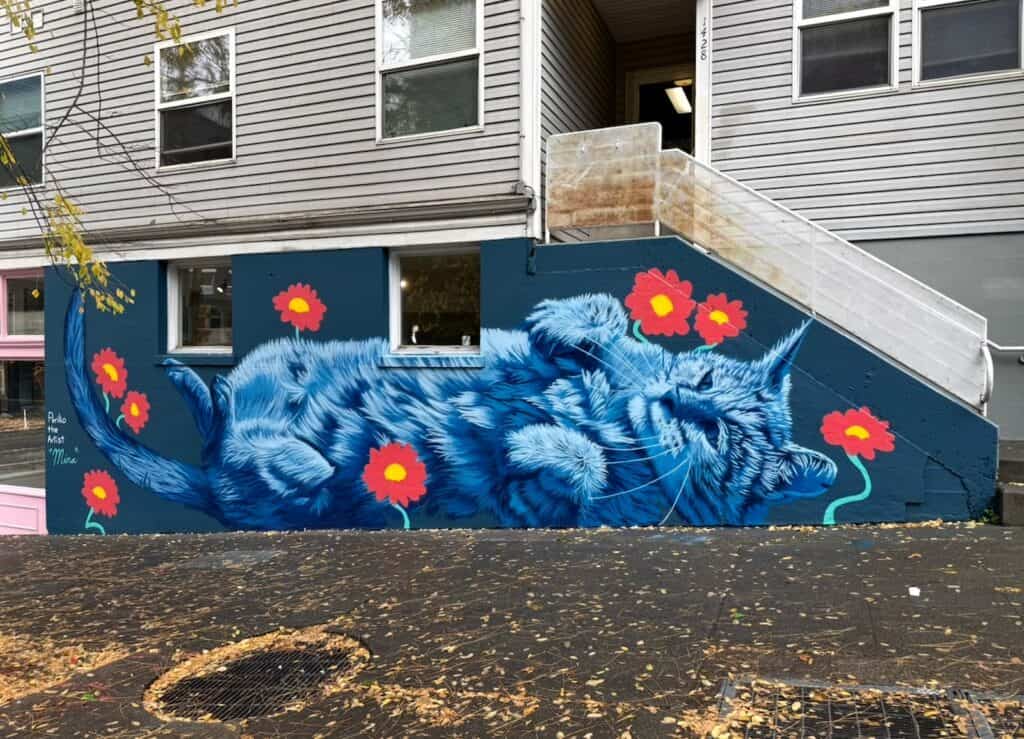 A Capitol Hill mural of a large, blue, fluffy cat lying on its back, surrounded by stylized red flowers.