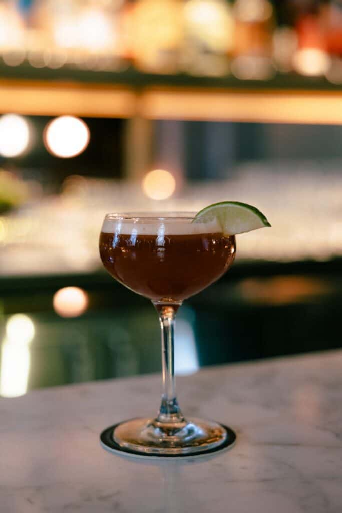A brown cocktail in a coupe glass garnished with a lime wedge rests on a marble bar top, perfect for winter drinks in Seattle.