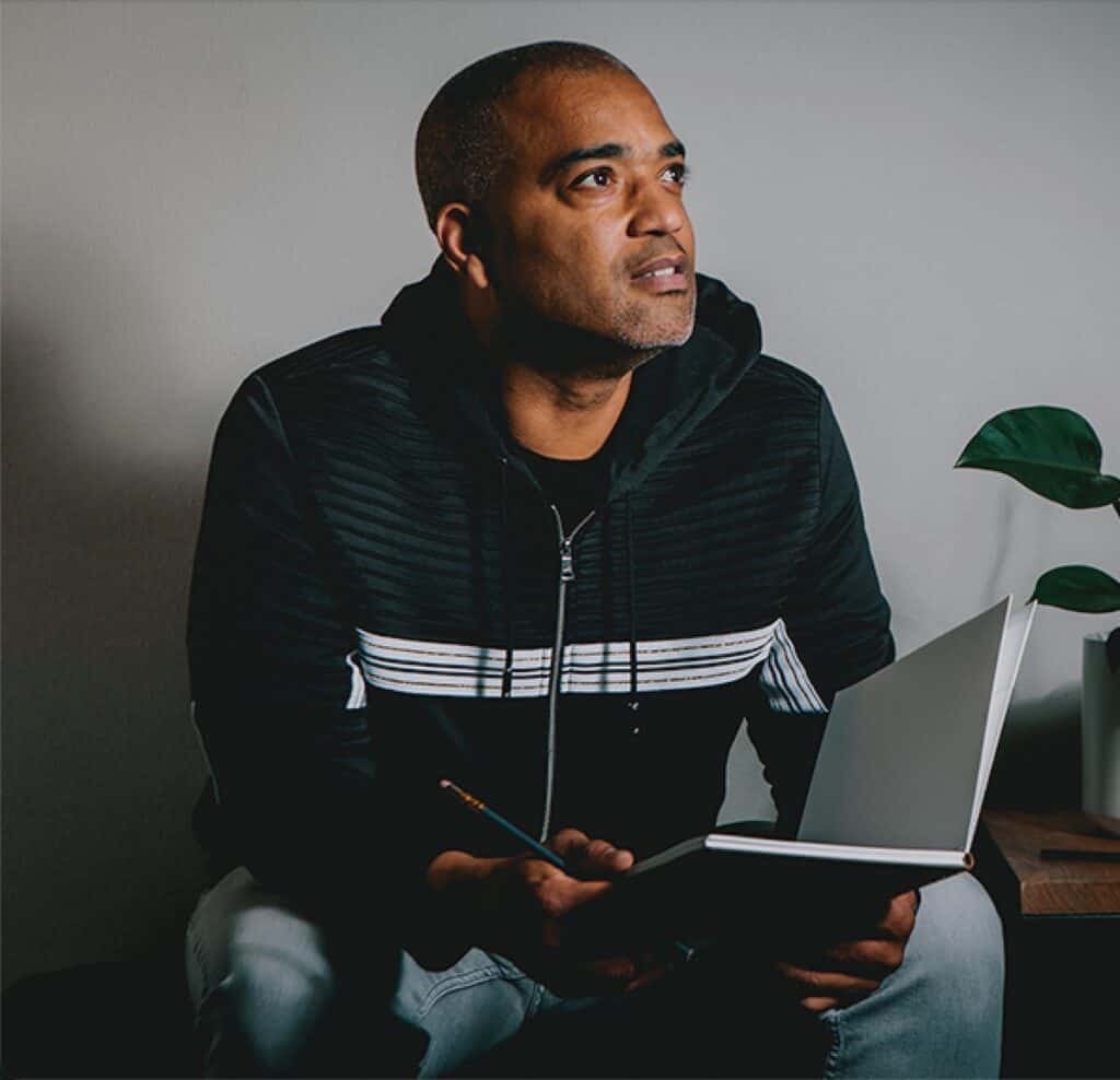 A man in a black zip-up hoodie sits holding a pencil and an open notebook, looking off to the side.