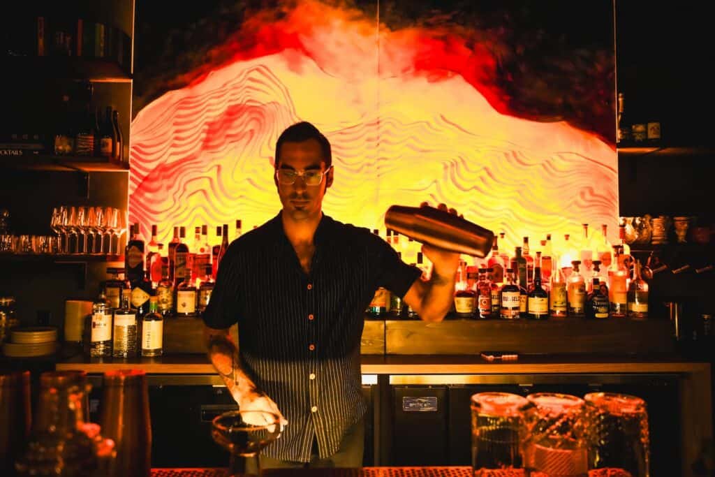 A bartender shaking a cocktail tin behind a bar with a backlit abstract art display.