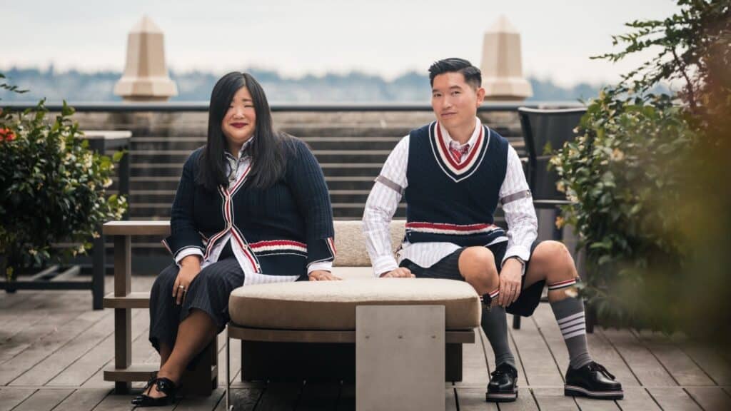 A man and a woman in matching navy sweater vests with red and white trim sit on a bench, a piece from the new furniture launch, as they look into a rearview mirror.