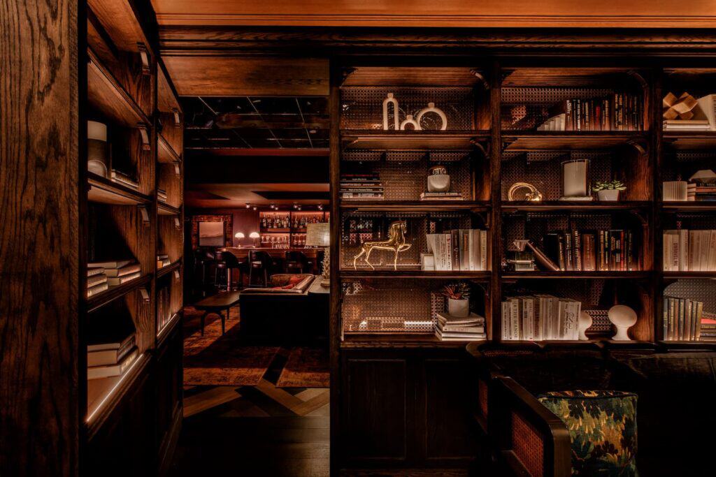 A dimly lit room with floor-to-ceiling wooden bookshelves, looking into a lounge area with a bar perfect for enjoying warm winter drinks.