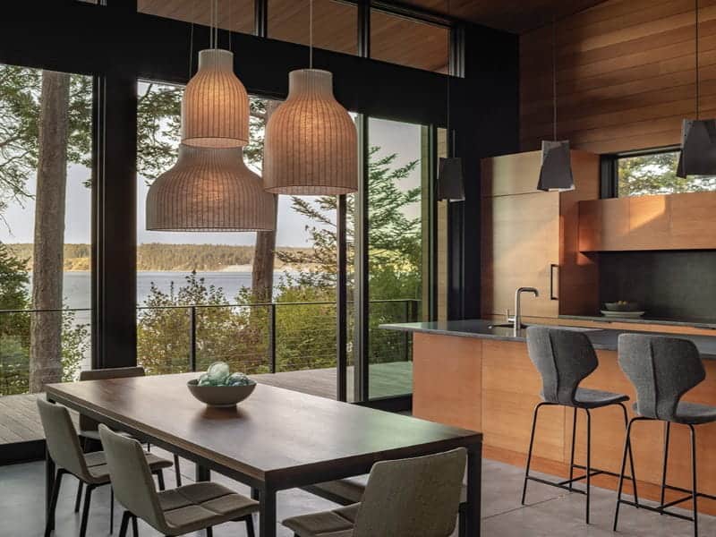 A Prentiss Balance Wickline modern kitchen and dining area features a wooden table, breakfast bar with stools, and three pendant lights, overlooking a lake through floor-to-ceiling windows.