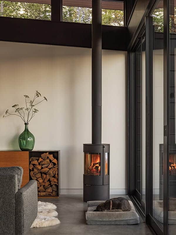 A modern, cylindrical wood-burning stove with a lit fire stands in a modern waterfront retreat, while a dog sleeps on a floor cushion nearby.