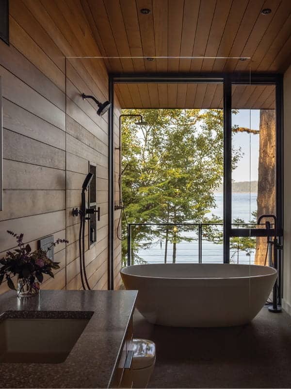 A modern San Juan Island bathroom featuring a freestanding white tub, wall-mounted shower fixtures, and a glass-walled view of the forest and water.
