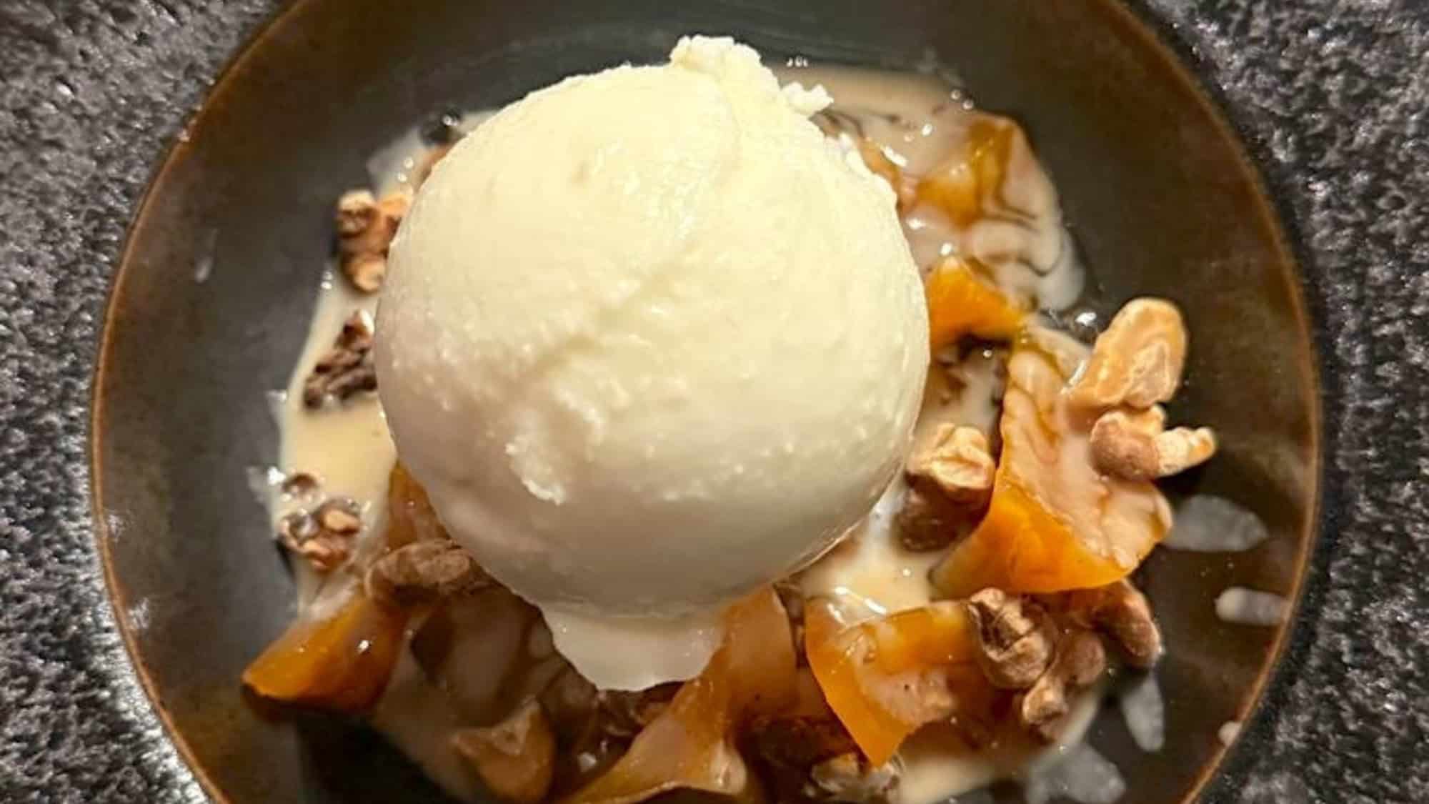 A scoop of vanilla ice cream sits atop walnuts and sliced caramelized fruit in a dark bowl, perfect for satisfying dessert cravings at Hey Bagel restaurant.