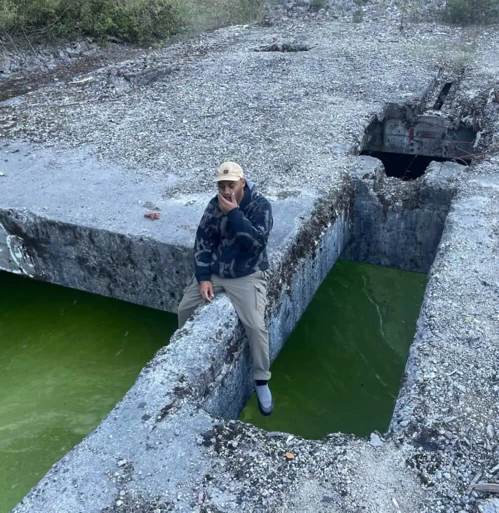 A person wearing a cap and hoodie sits on a concrete ledge over a rectangular pool of bright green water.