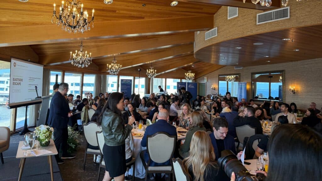 A speaker stands with a microphone addressing a crowded brunch event in a large room with vaulted wood ceilings and multiple tables of guests.