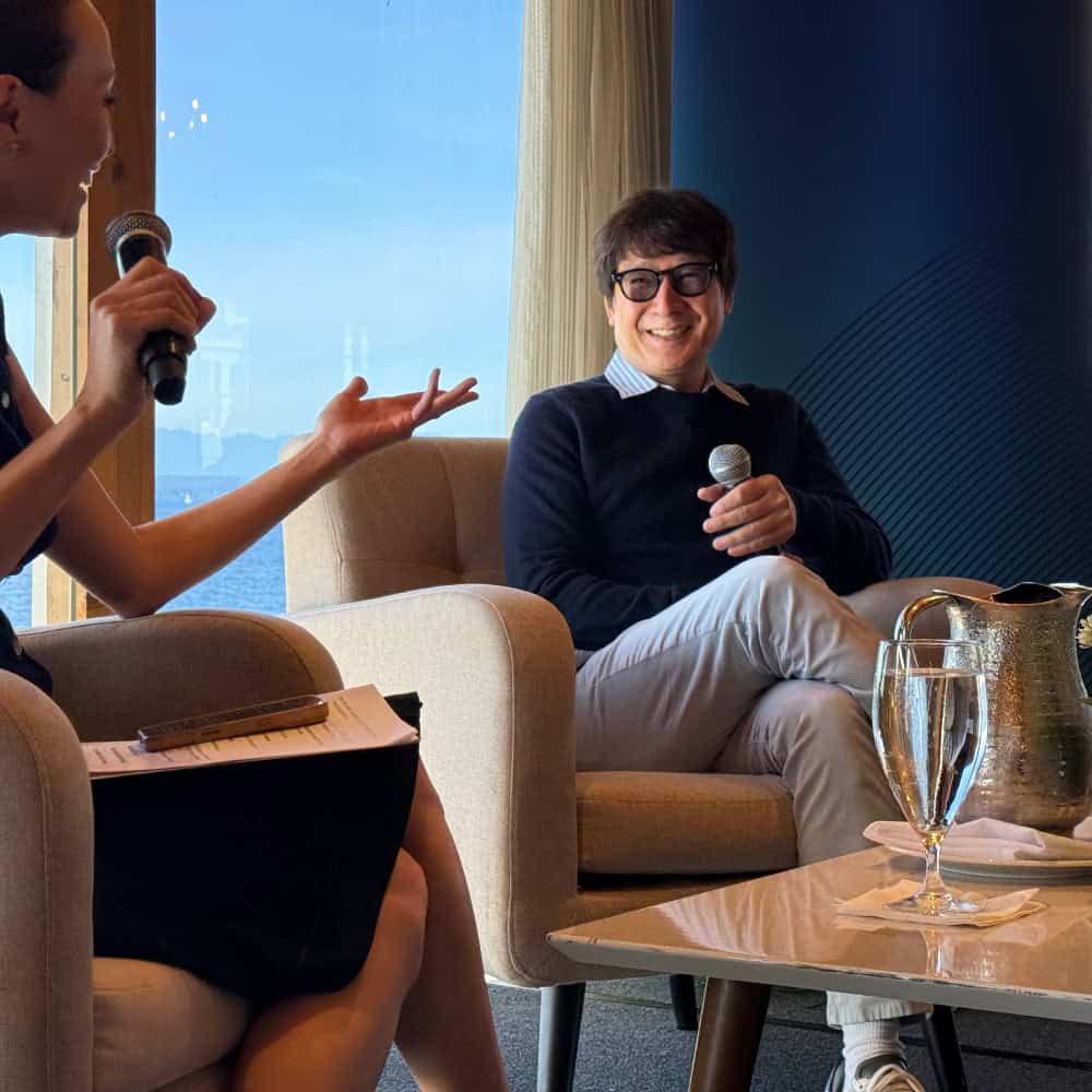 A person interviewing a man in glasses who is holding a microphone and smiling while seated in an armchair.