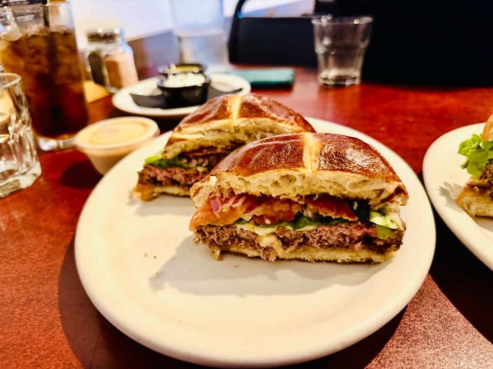 A hamburger on a toasted pretzel bun, cut in half on a white plate and ready for our counter culture food guide.