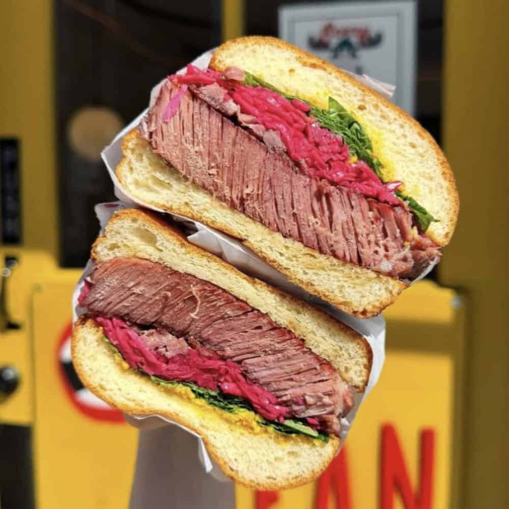 Two halves of a MEAN pastrami sandwich on sliced bread with pink kraut and greens.